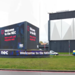 Digital welcome sign reads, "Welcome to the NEC. Have a safe and enjoyable visit." It's mounted on a grassy area with a large building and parked vehicles in the background.