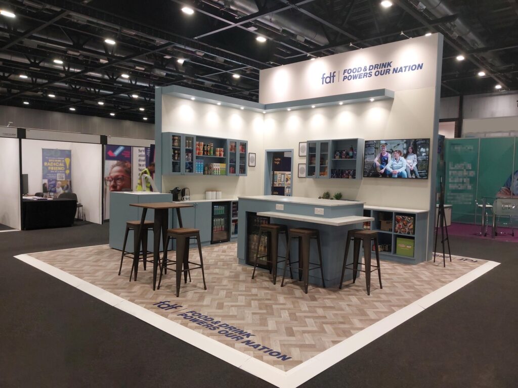 Exhibition booth with signage reading "Food & Drink Powers Our Nation," featuring shelves, a central counter with stools, and a TV displaying a video.