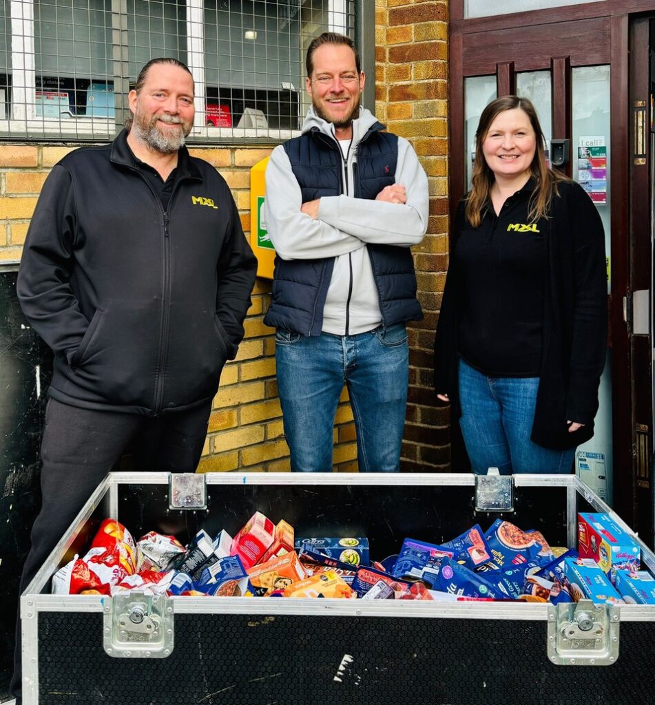 Three people stand smiling behind a large box filled with various packaged food items, in front of a brick wall.